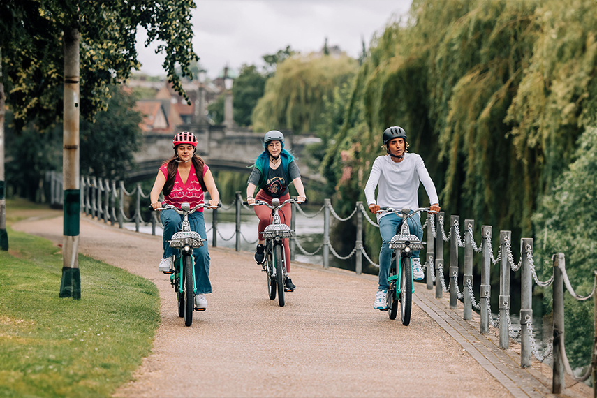 Three people are riding bicycles along a riverside pathway bordered by trees and a metal chain fence. The individuals are wearing helmets, and they appear to be enjoying the scenic ride on a cloudy day. A bridge and more greenery are visible in the background.