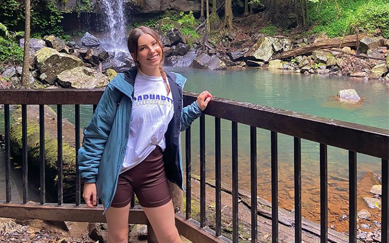 A woman is standing in front of a wooden railing with a tranquil waterfall and small pond in the background. She is wearing a blue jacket, white T-shirt, and brown shorts, smiling at the camera. Surrounding her are lush green trees and rocks.