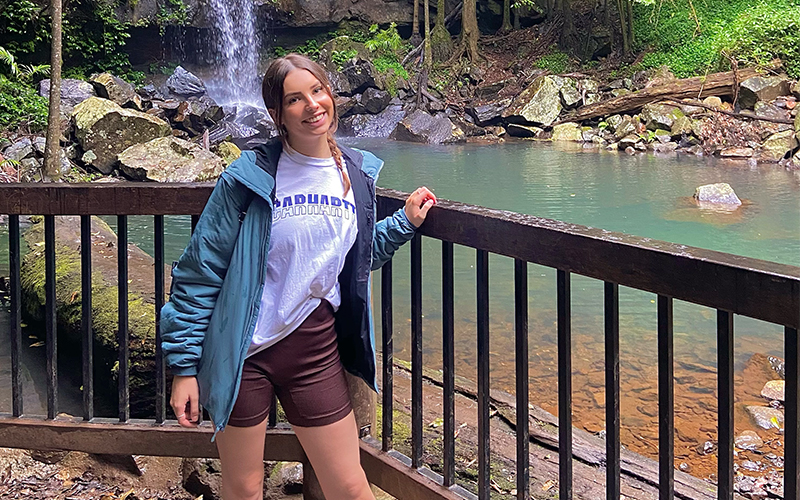 A woman is standing in front of a wooden railing with a tranquil waterfall and small pond in the background. She is wearing a blue jacket, white T-shirt, and brown shorts, smiling at the camera. Surrounding her are lush green trees and rocks.