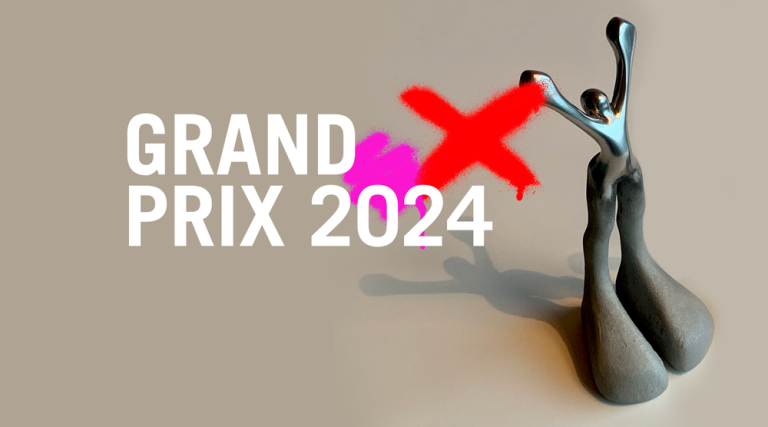 A gray metallic sculpture stands on a smooth, gray surface. To the left, bold white text reads "GRAND PRIX 2024" with "JUX" partly crossed out in bright red and pink paint strokes, partially obscuring the text. The background is light gray.