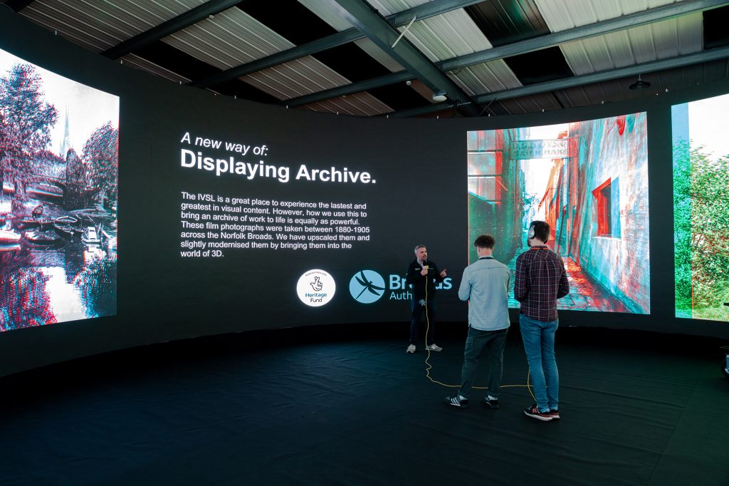 Three people stand in front of a large, curved screen displaying digital visualisations and text about displaying archive files using VR technology. The screen showcases both monochrome and colourful images, including landscapes and buildings, under an arched roof.