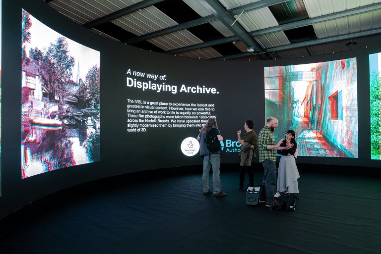 A group of people standing and talking in front of a large curved digital display. The display shows text titled, "A new way of: Displaying Archive," along with several colourful digital images. The setting is a modern exhibition space with a high ceiling.