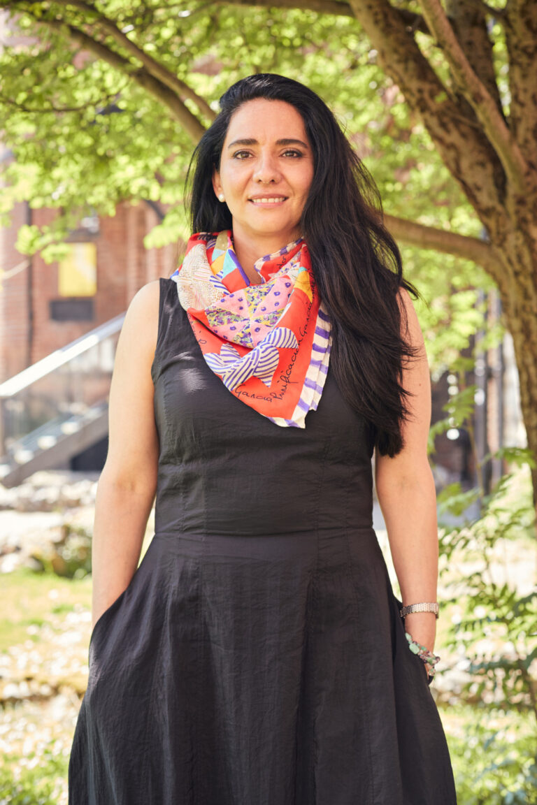 A person with long dark hair stands outdoors in front of a tree, wearing a sleeveless black dress and a colourful, patterned scarf. They are smiling and have their hands in their dress pockets. The background features green foliage and a blurred building.