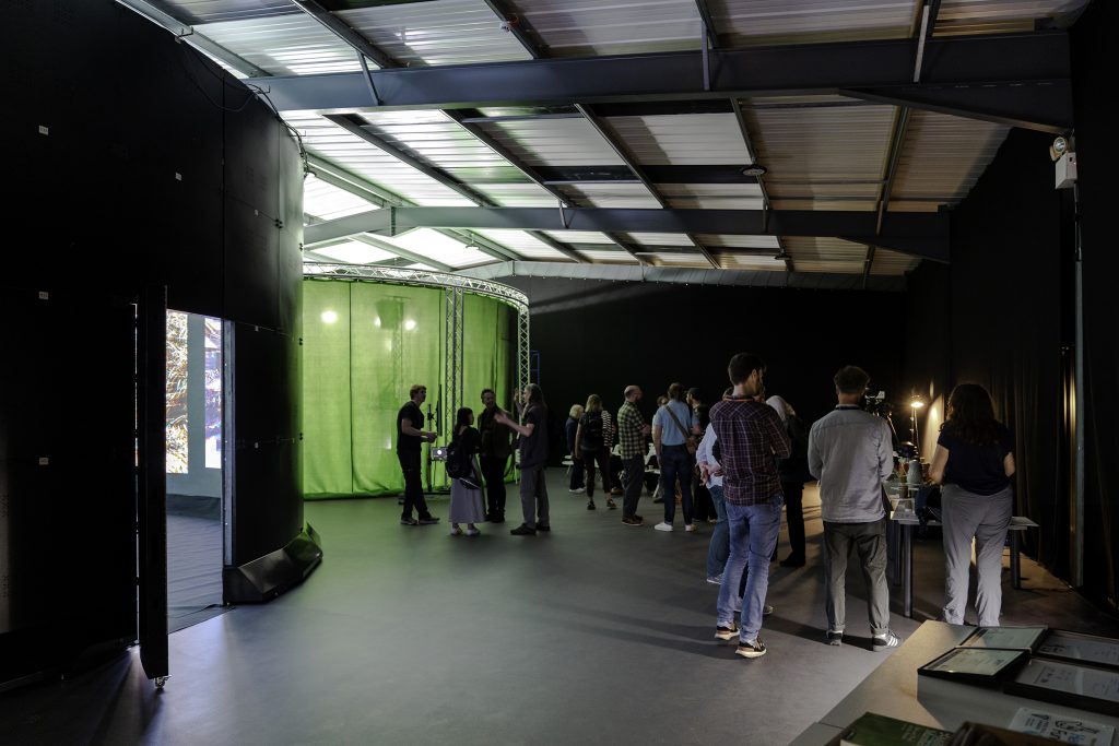 A group of people are gathered in an indoor space with a high ceiling. To the left, there's a green screen setup with lights. To the right, others are standing near tables with equipment. The room has a modern, industrial feel with metal beams and concrete floors.