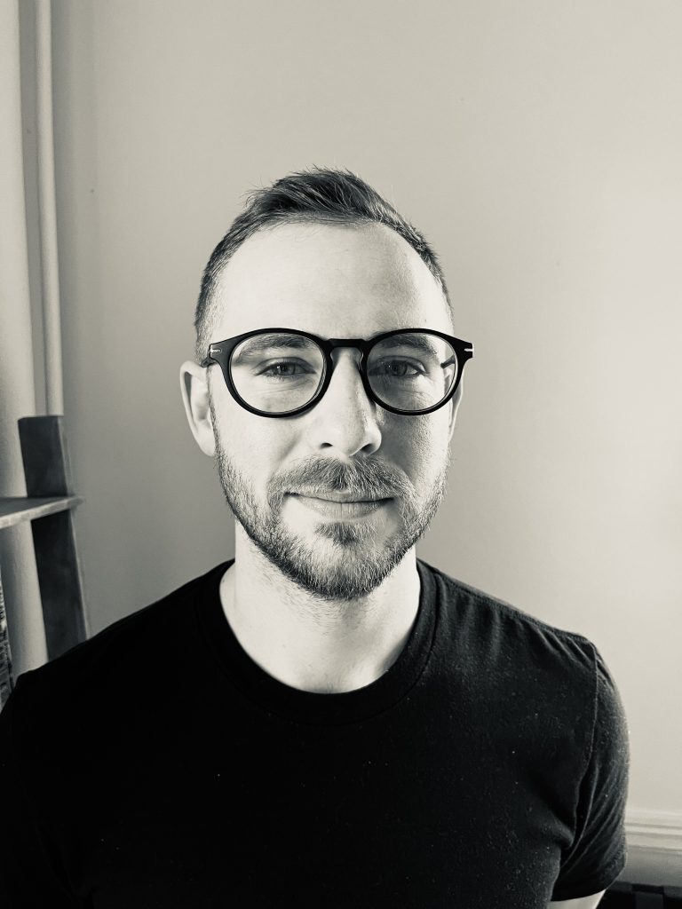 Black and white portrait of a person with short hair, wearing glasses and a black shirt, looking directly at the camera with a slight smile. The background is simple, possibly a wall, with part of a wooden structure visible.