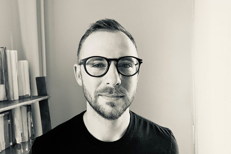 Black and white portrait of a person with short hair, wearing glasses and a black shirt, looking directly at the camera with a slight smile. The background is simple, possibly a wall, with part of a wooden structure visible.