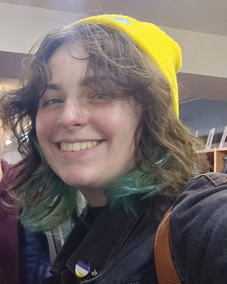 A person with shoulder-length hair, smiling, wearing a bright yellow beanie and a dark jacket. Their hair has green tips, and shelves with small items are visible in the background.