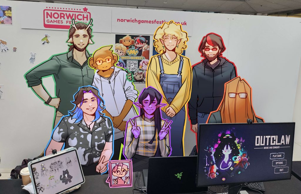 A group of colourfully outlined characters stands behind a table with monitors displaying game graphics. The background features Norwich Games Festival signage.