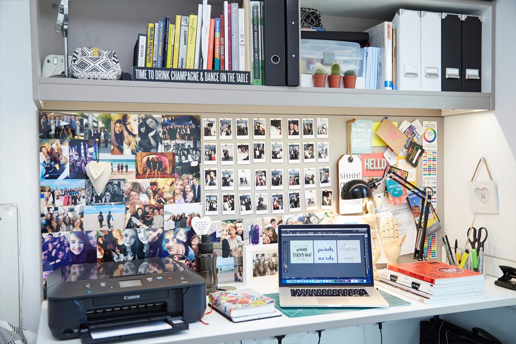 A cluttered desk setup with a laptop surrounded by books, a Canon printer, a corkboard filled with photos, notes, shelves with more photos, plants, office supplies, and decorative items. Shelves above hold stationery, storage boxes, and books.