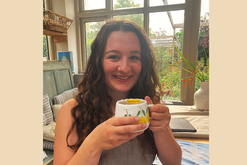 A woman with long hair smiles while holding a white mug with a yellow pattern. She is sitting in a cozy, sunlit room with large windows and various potted plants in the background.