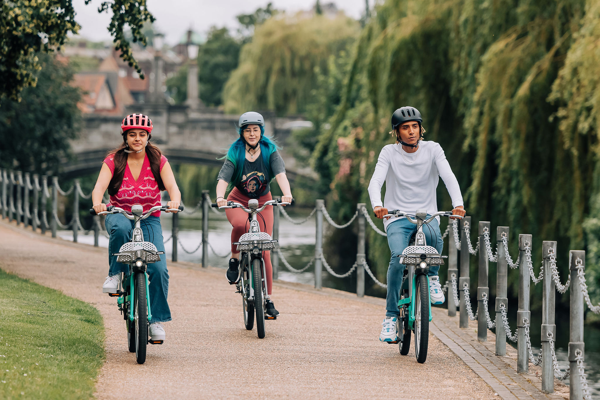 Three people are riding bicycles along a riverside pathway bordered by trees and a metal chain fence. The individuals are wearing helmets, and they appear to be enjoying the scenic ride on a cloudy day. A bridge and more greenery are visible in the background.