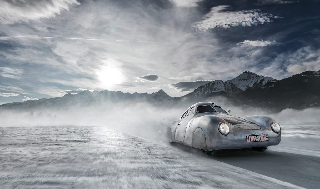 A silver vintage car drives at high speed across a snowy landscape with mist trailing behind. The backdrop features majestic, snow-covered mountain peaks under a partly cloudy sky with the sun shining through. The car has a red license plate.