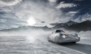 A silver vintage car drives at high speed across a snowy landscape with mist trailing behind. The backdrop features majestic, snow-covered mountain peaks under a partly cloudy sky with the sun shining through. The car has a red license plate.