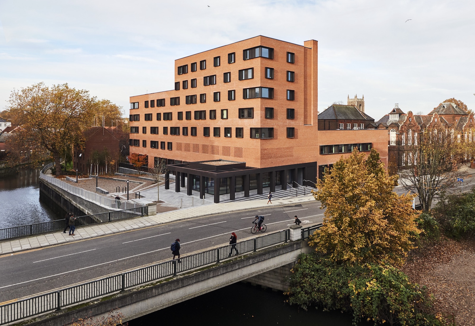 An angular brick building with multiple windows stands next to a river, featuring a modern entrance area. People are walking and cycling on the adjacent bridge. Autumn trees and historic buildings create a picturesque background.