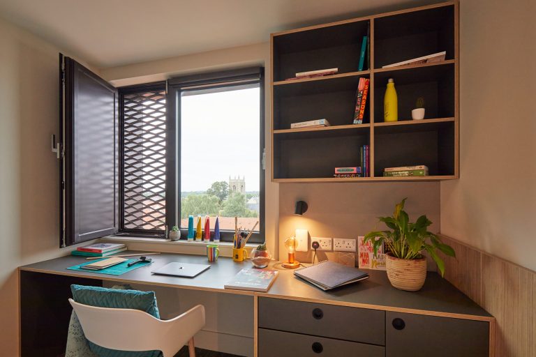 A warm and neatly organised desk space featuring a desk with a laptop, books, stationery, a potted plant, and a lamp. Above the desk are open shelves with more books and decor items. A window with a view of trees and a church steeple adds natural light to the room.