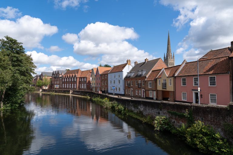 A row of colourful houses with steep roofs lines a river under a bright blue sky with fluffy clouds. The calm water reflects the buildings and sky.