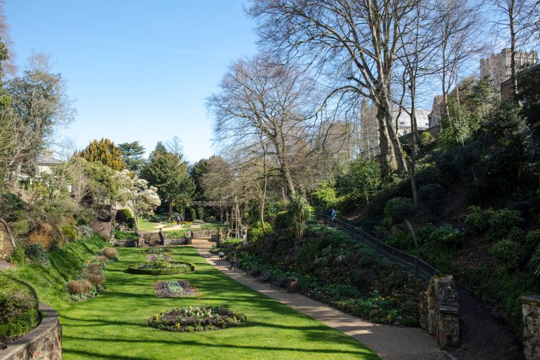 A well-maintained garden with symmetrically arranged flower beds and manicured lawns. The garden is bordered by tall trees and shrubbery. A pathway winds through the greenery and leads towards an archway. The sky above is clear and bright blue.