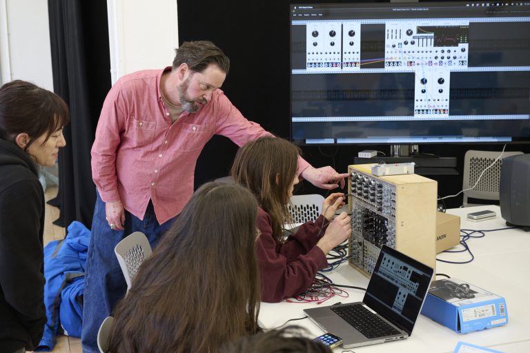 A man in a red shirt instructs two individuals on how to operate electronic music equipment with a computer screen displaying related software in the background. The setup includes laptops and various electronic devices.