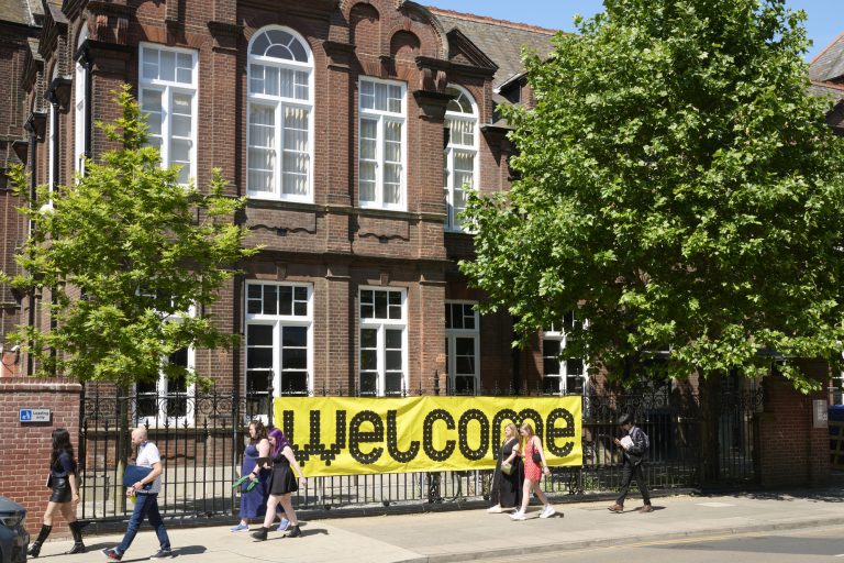 A group of people walking in front of a large brick building with tall windows. The building has a large yellow banner hanging on the fence that reads "Welcome." Trees are visible, providing shade along the street.