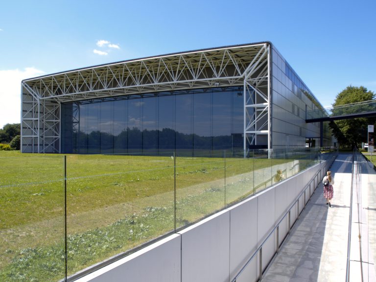 A modern, rectangular building with a glass facade and a metal framework. There is a grassy field in front of it. The sky is clear and blue.