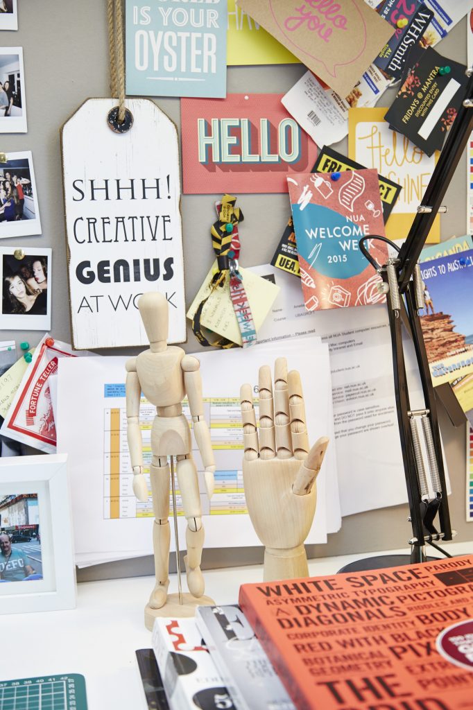 A cluttered desk with a bulletin board filled with colourful notes and signs, such as "SHHH! Creative Genius at Work," and "Hello." A wooden mannequin and a hand model are also on the desk, along with various papers and photos.