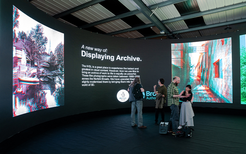 A group of people standing and talking in front of a large curved digital display. The display shows text titled, "A new way of: Displaying Archive," along with several colourful digital images. The setting is a modern exhibition space with a high ceiling.