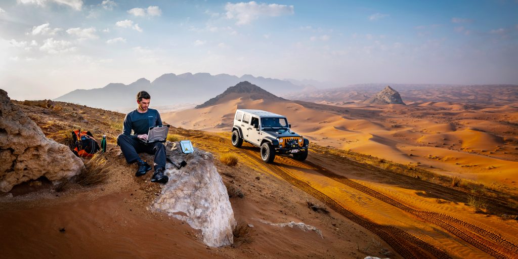 A person sits on a rock in a desert landscape, using a laptop, with a backpack and tablet nearby. An off-road vehicle is parked on a sandy track beside them. Mountainous terrain is visible in the distance under a partially cloudy sky.