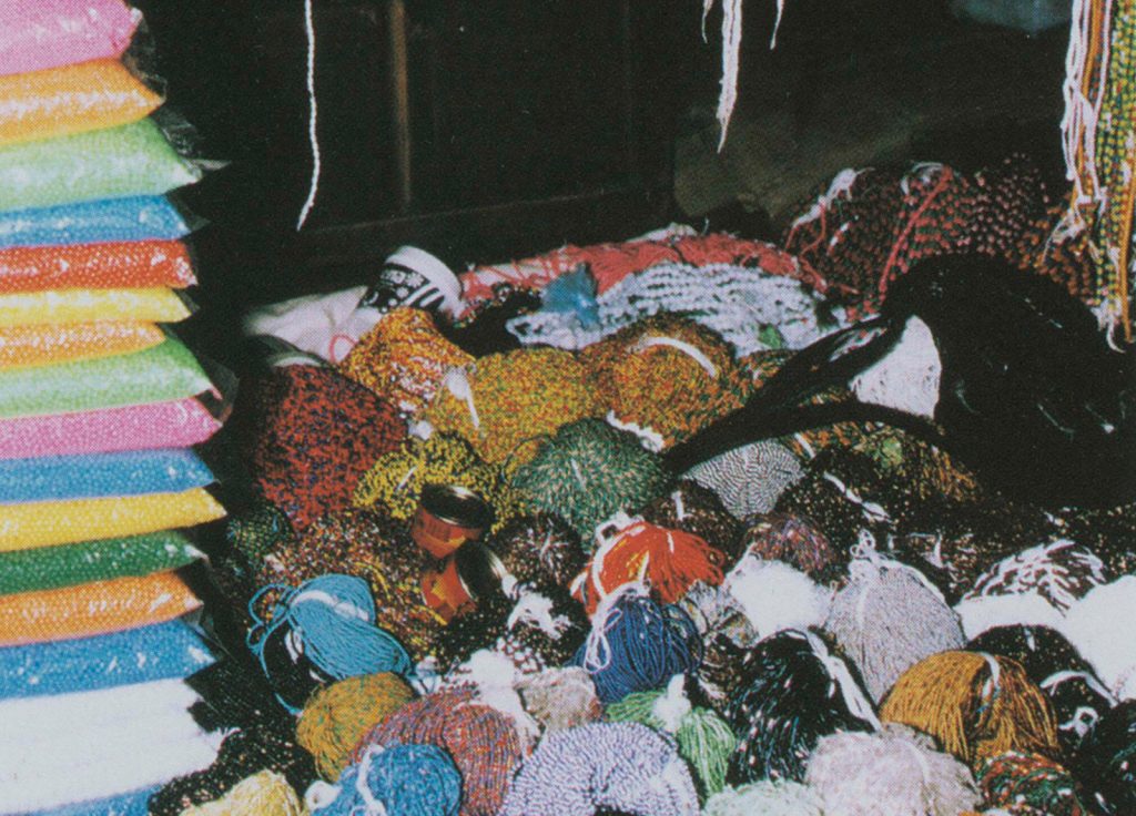 A chaotic array of colourful beads and strings fills the surface, creating a vibrant but cluttered scene. To the left, there are stacked bags in various bright colors. Among the beads and strings, there are some unidentifiable objects and partially visible items.