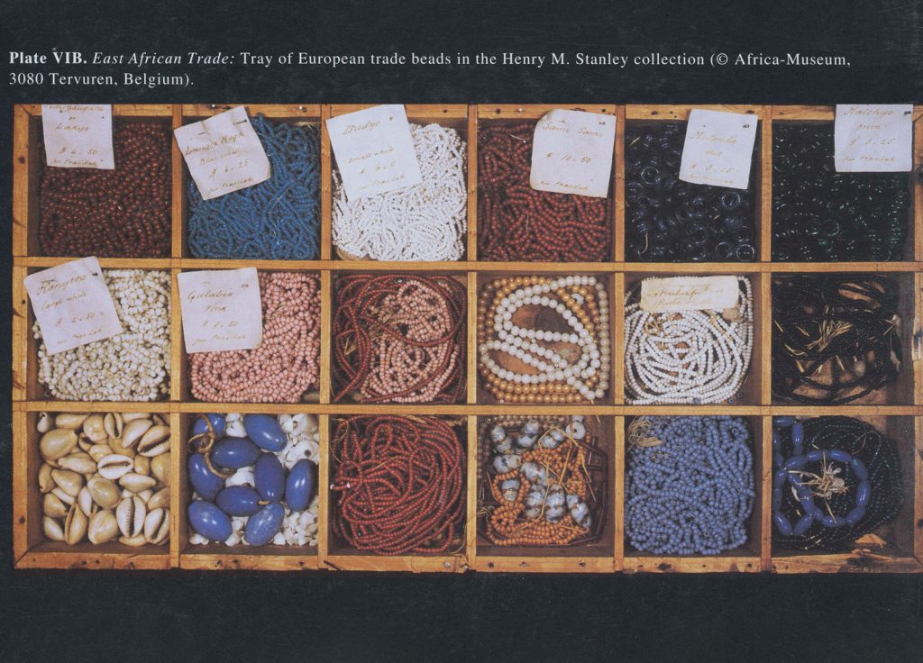 A wooden tray subdivided into 18 compartments, displaying various trade beads of different colors and shapes. Some compartments have handwritten labels. The tray is part of the Henry M. Stanley collection at the Africa-Museum in Tervuren, Belgium.