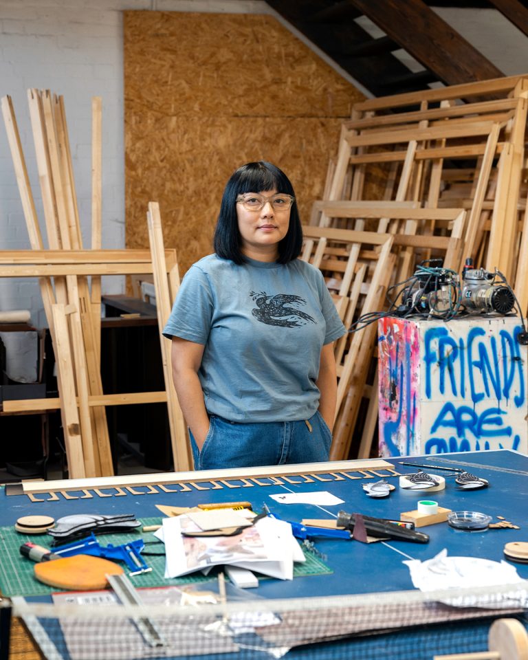 A person with long black hair and glasses stands in an art studio. They wear a blue t-shirt and jeans. Various art supplies and tools are on a table, and wooden frames are stacked in the background.