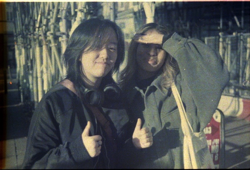 Two people stand outdoors. The person on the left is smiling and giving a thumbs-up. The person on the right shades their eyes with one hand. Both are wearing jackets and appear to be in a sunny urban setting.