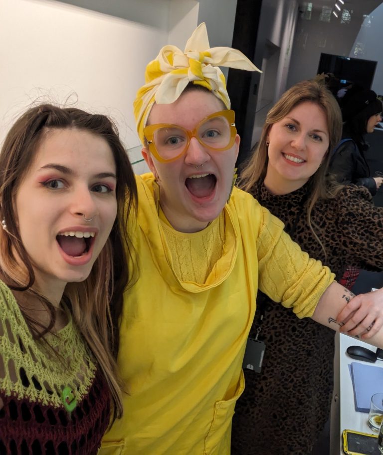 Three people smiling for a selfie indoors. The person on the left has long hair and a knitted top, the middle person wears a bright yellow outfit with a headscarf and glasses, and the person on the right is in a patterned dress.