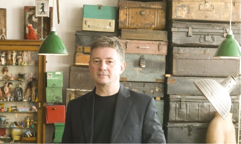 A man in a black pinstripe suit stands in front of a stack of vintage suitcases and various storage boxes. There are two green hanging lamps overhead. To his left, there is a glass cabinet displaying figurines and small items.