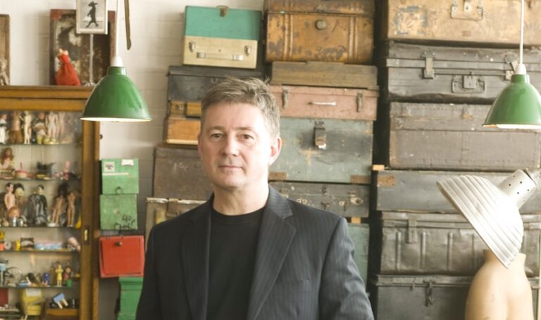 A man in a black pinstripe suit stands in front of a stack of vintage suitcases and various storage boxes. There are two green hanging lamps overhead. To his left, there is a glass cabinet displaying figurines and small items.