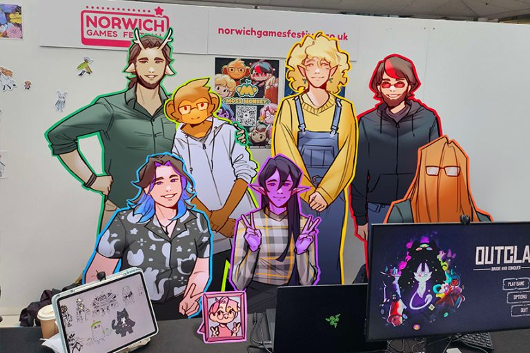 A group of colourfully outlined characters stands behind a table with monitors displaying game graphics. The background features Norwich Games Festival signage.