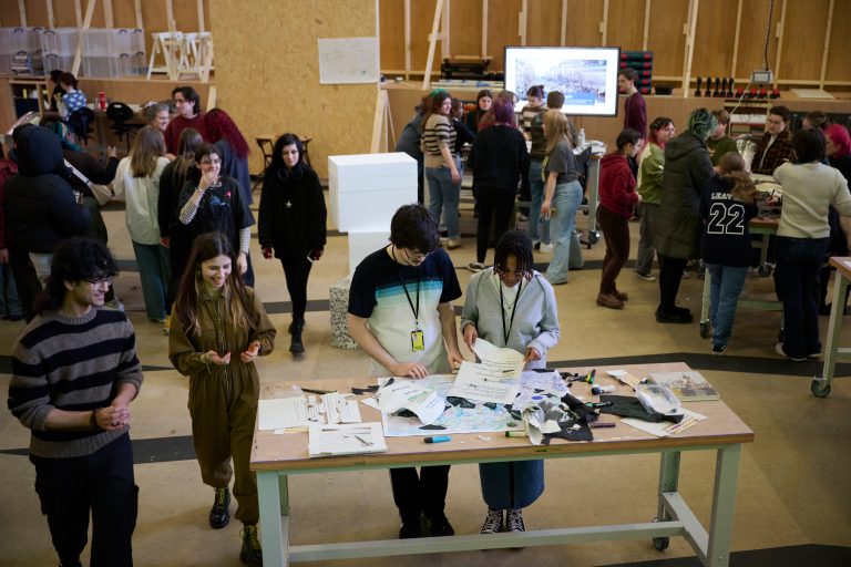 A busy workshop scene shows several people working at tables covered with papers and materials. Some groups are standing and discussing, while others are focused on tasks. The atmosphere is collaborative.