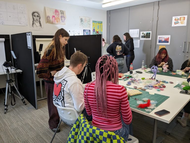 A group of people engaged in crafting activities around a table covered with colourful materials. Artworks and drawings adorn the walls.