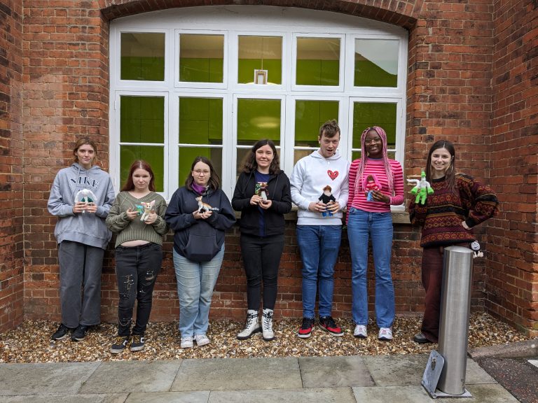 Seven people stand in front of a brick building with a white-framed window. They each hold small felted figures. The group is casually dressed in jackets and sweaters.