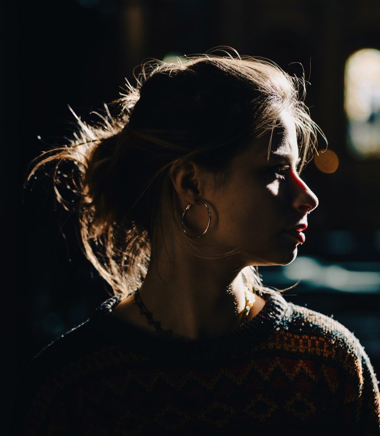 A person in profile with brown hair in a bun, wearing hoop earrings and a patterned sweater, is illuminated by dramatic side lighting in a dark setting.