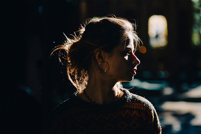 A person in profile with brown hair in a bun, wearing hoop earrings and a patterned sweater, is illuminated by dramatic side lighting in a dark setting.