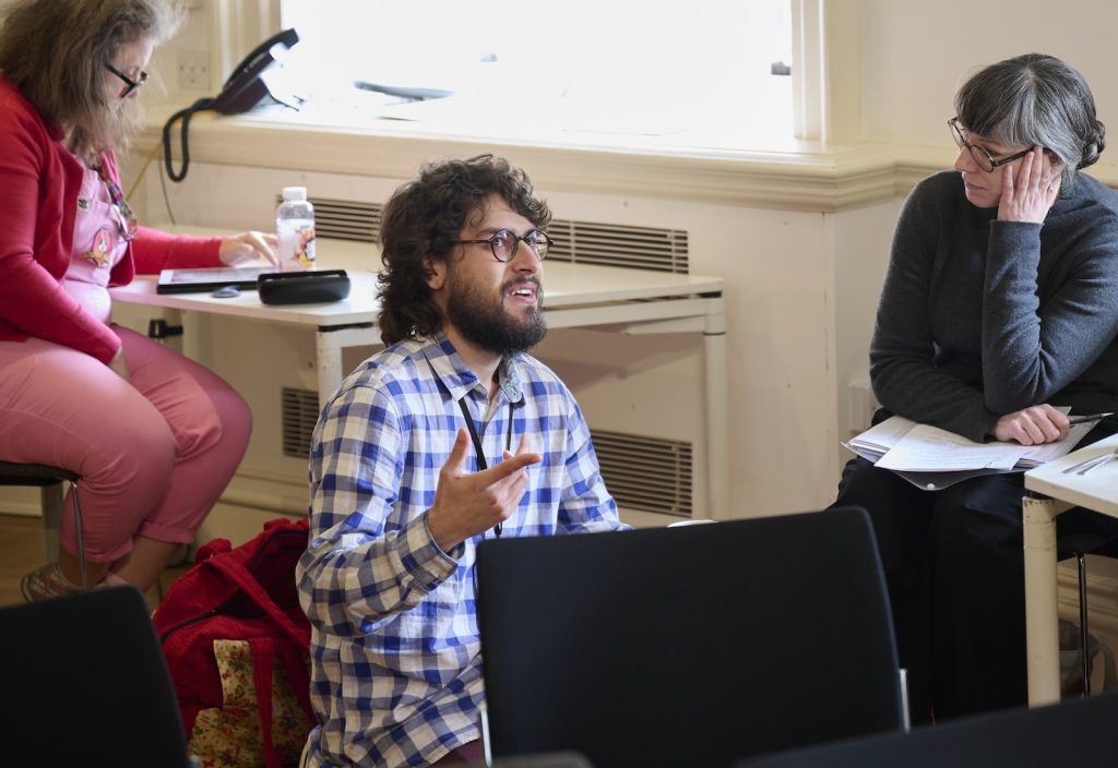 A man with glasses and a beard gestures passionately while sitting in a classroom. A woman in a grey sweater and another in pink attire sit nearby, listening intently. Papers and notebooks scatter across the desks.