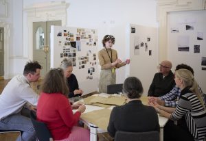 A group of people sits around a table, applying insights from research as they actively listen to someone standing and speaking. The room's white walls are adorned with various photos and artworks, suggesting an inspiring environment for this creative workshop or meeting focused on a degree pursuit.