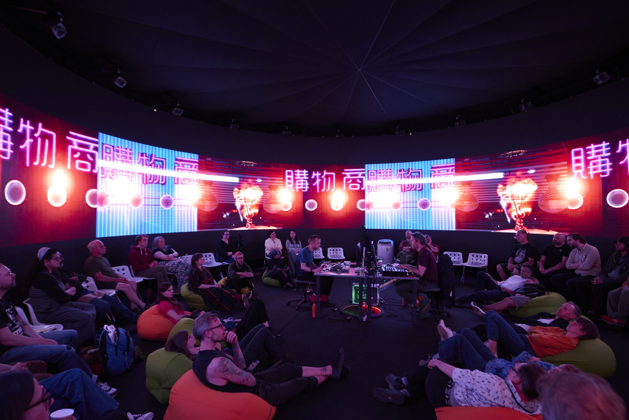 People sit and lounge on bean bags and chairs in a dark circular room with neon pink and blue digital screens displaying Japanese characters and abstract graphics around the walls.