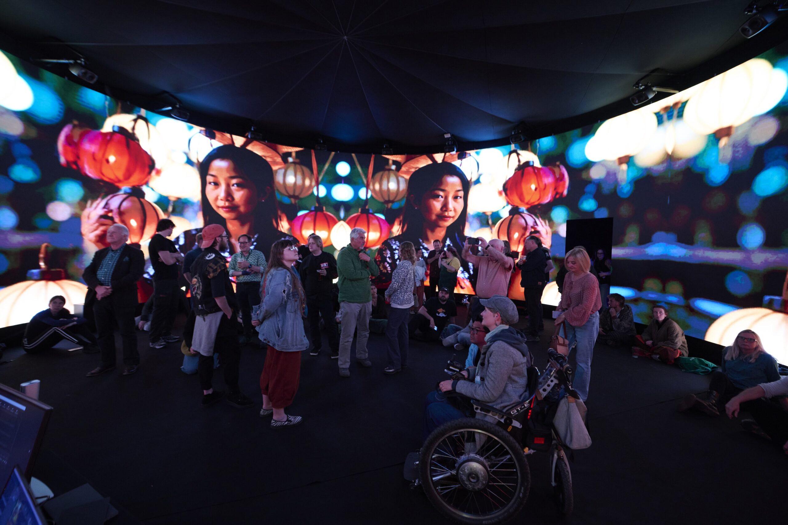 A diverse group of people, including a person in a wheelchair, stand inside a dome with large, colorful digital screens showing a woman's face and lanterns, creating an immersive, vibrant atmosphere.