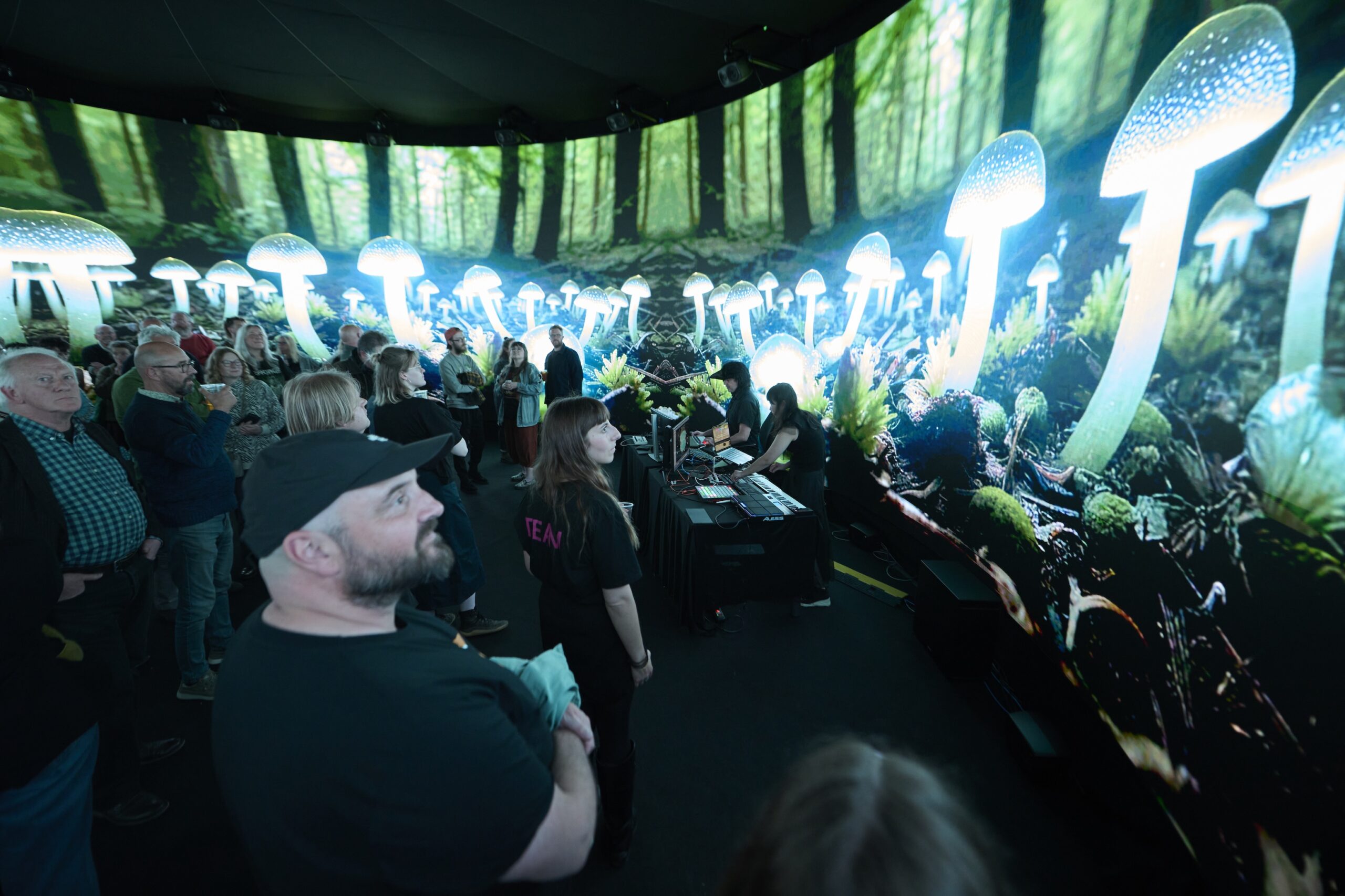 A group of people stand inside an immersive exhibit with large, curved screens displaying glowing, oversized mushrooms and a lush forest scene, creating a surreal, interactive environment.