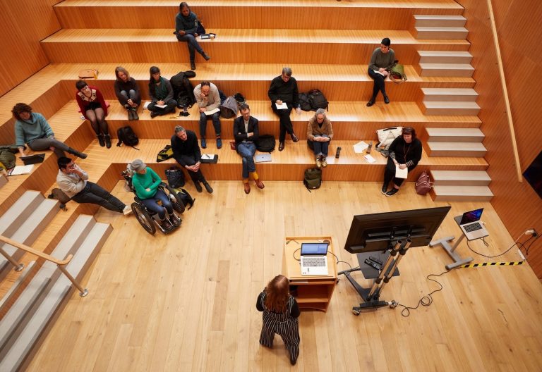 A person stands at a podium with a computer and screen, presenting their research to an audience seated on tiered wooden steps in an auditorium. The group listens attentively, taking notes, eager to apply the insights to their degree studies.