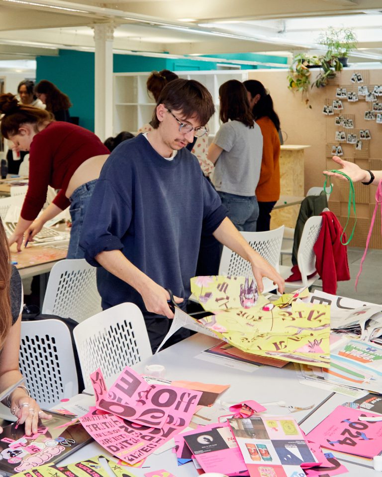 A person in glasses and a dark shirt uses scissors to cut colourful paper with printed text and graphics at a busy workshop table. Various papers and art supplies are scattered around. People in the background work on similar projects.