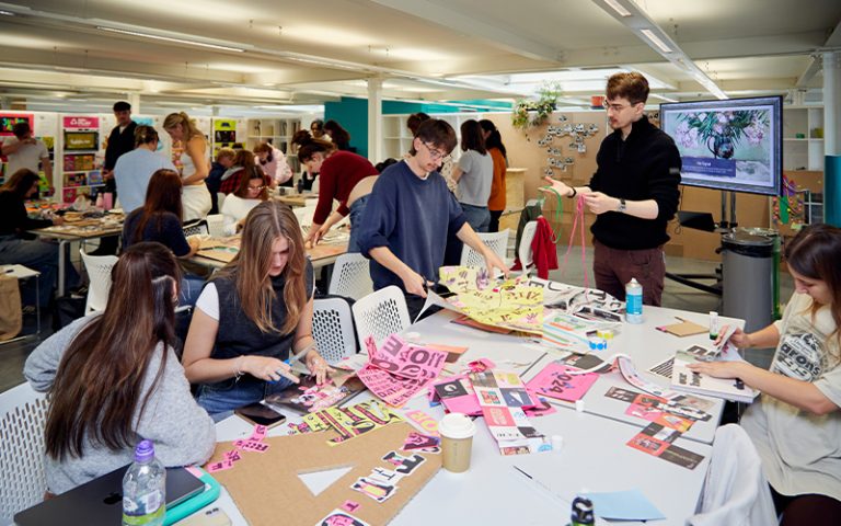 A lively classroom setting with students engaged in a creative workshop. They are gathered around tables covered with colourful papers, magazines, and art supplies, collaborating on design projects. A screen displays a digital design in the background.