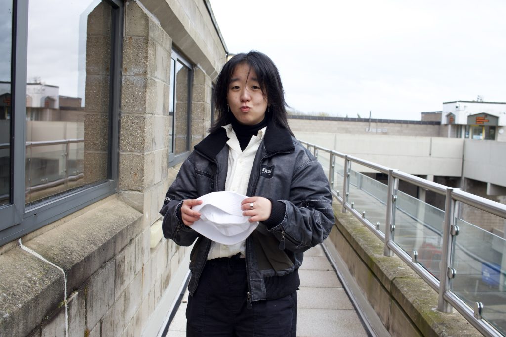 A person with long dark hair stands on an outdoor walkway, holding a white hat. They are wearing a black jacket over a white shirt, with a neutral expression. The background shows a building with large windows and a cloudy sky.