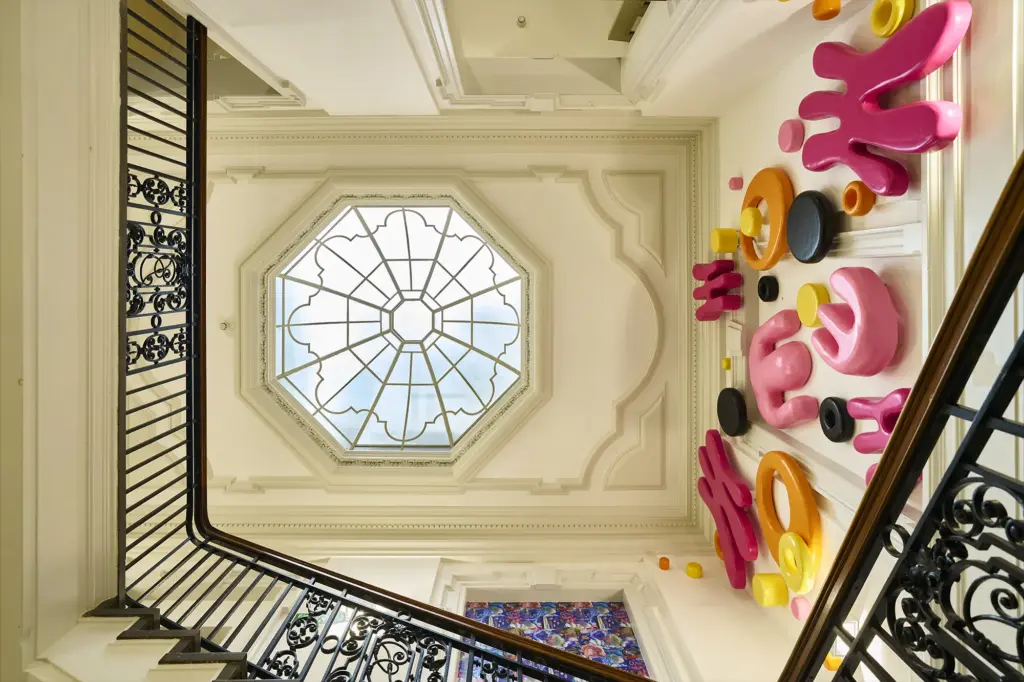 View looking up a stairwell with a large octagonal skylight and white decorative molding. Bright pink, orange, yellow, and black abstract wall sculptures are mounted on the right-hand wall. Black railings frame the image.